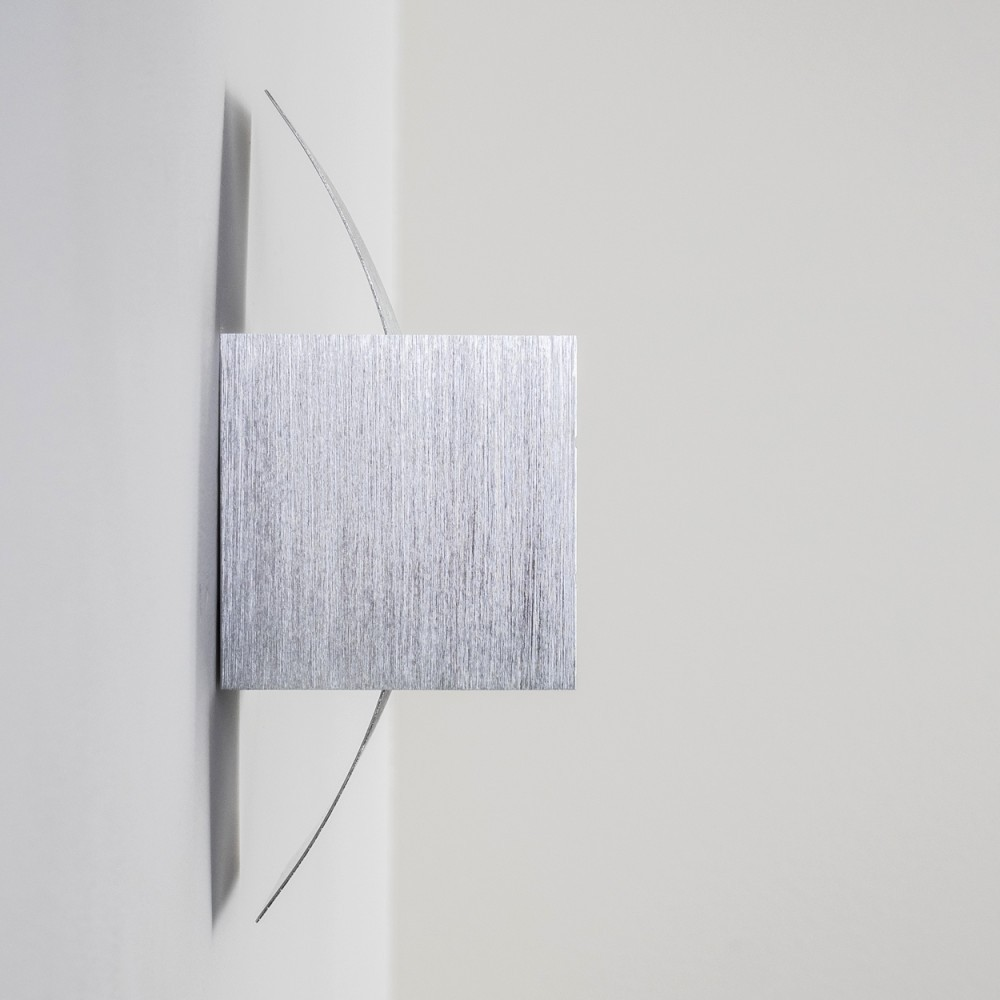 Moderne silberne Wandskulptur. Ein quadratischer Metallblock mit vertikalen Linien und gebogenen Metallstreifen an einer weißen Wand.