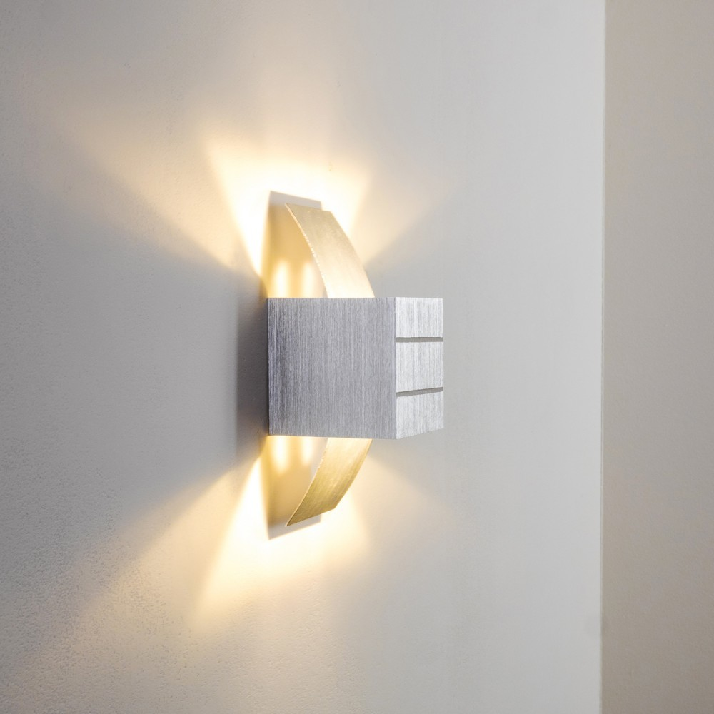 Eine moderne silberne Wandlampe mit goldenen Akzenten an einer weißen Wand. Die Lampe strahlt warmes Licht aus und wirft Schatten.