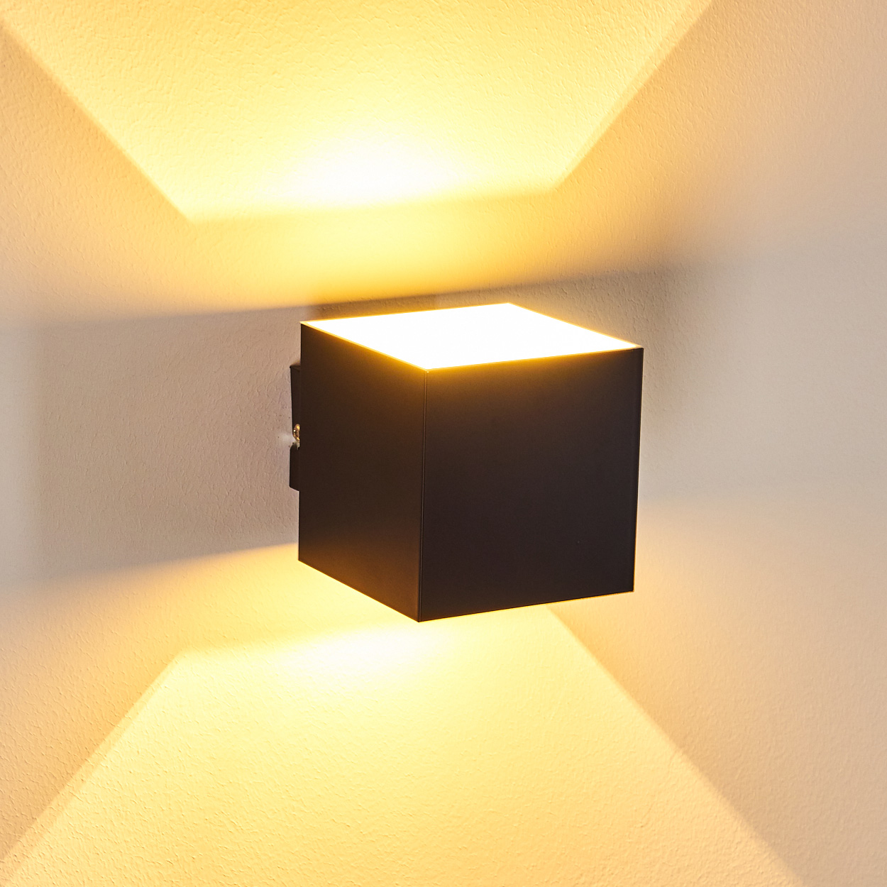 Quadratische, schwarze Leuchte an strukturierter Wand. Licht scheint nach oben und unten und erzeugt warmen Schein und Schatten.