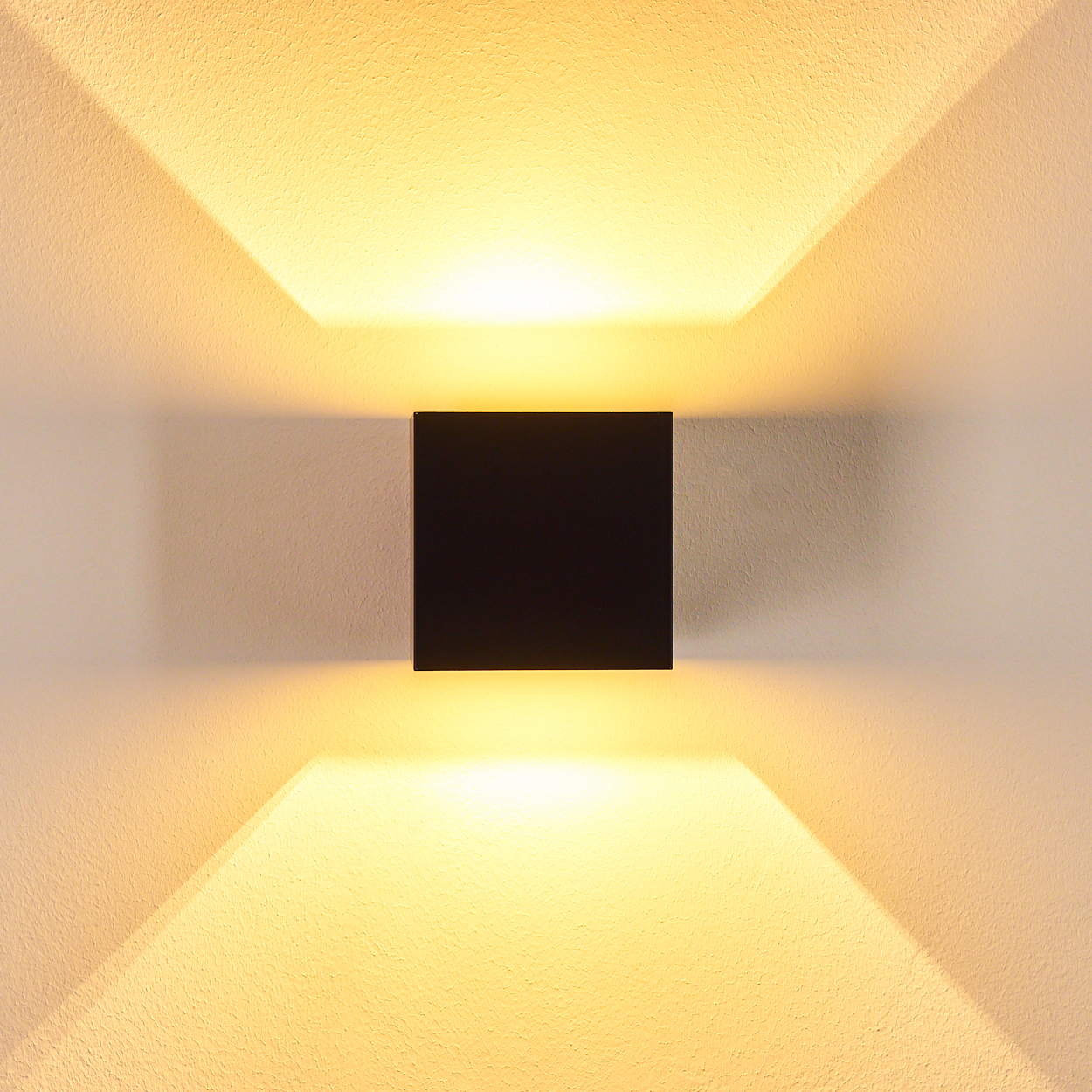 Eine quadratische, schwarze Wandleuchte ist zentriert. Das Licht strahlt nach oben und unten und erzeugt ein symmetrisches, beleuchtetes Muster.