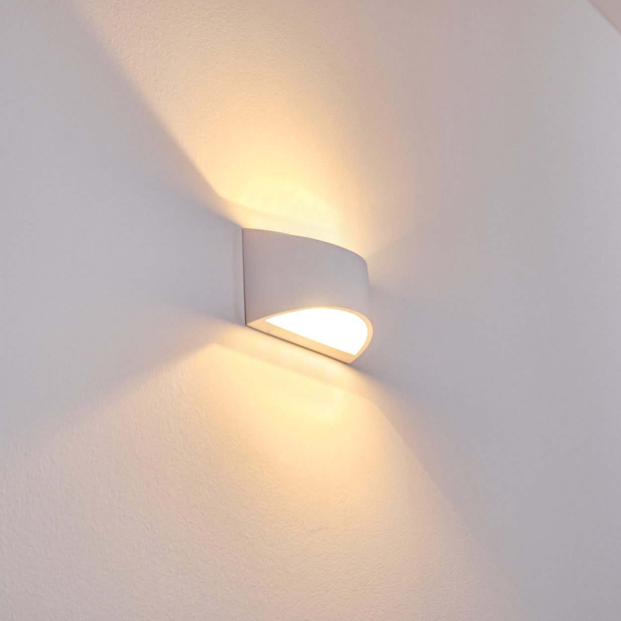Eine weiße, an der Wand befestigte Leuchte mit gebogenem Design. Sie strahlt warmes Licht aus und wirft Schatten auf die weiße Wand.
