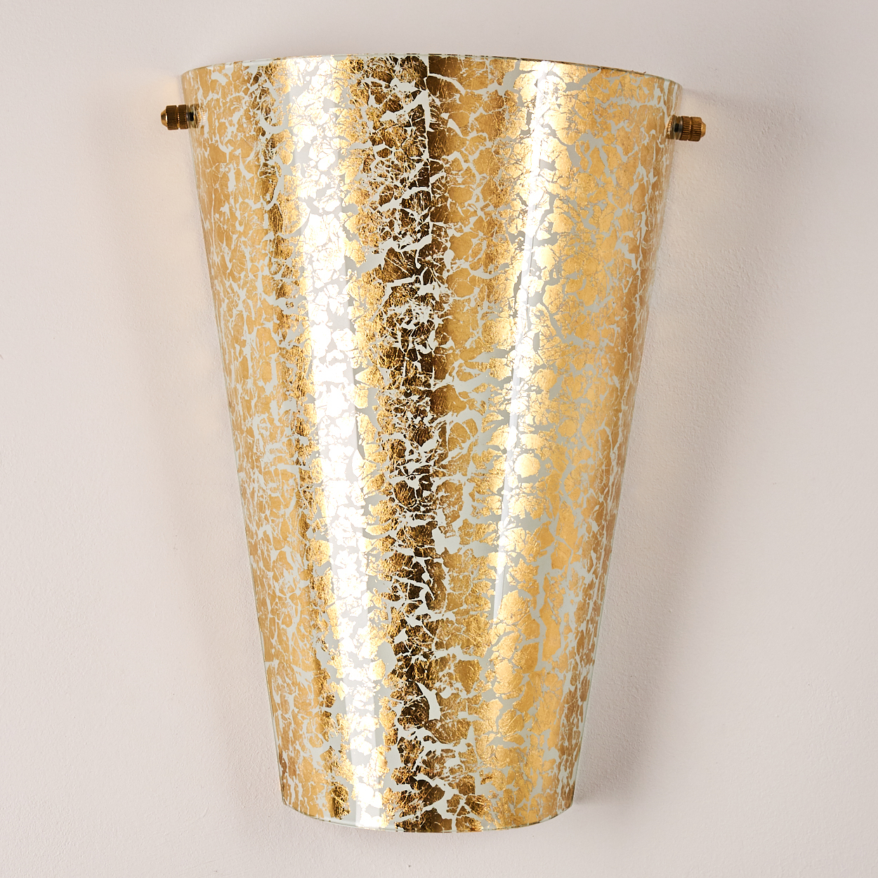 Eine goldene Wandleuchte mit Rissmuster, an einer weißen Wand befestigt. Zwei kleine Stifte halten die Leuchte fest.