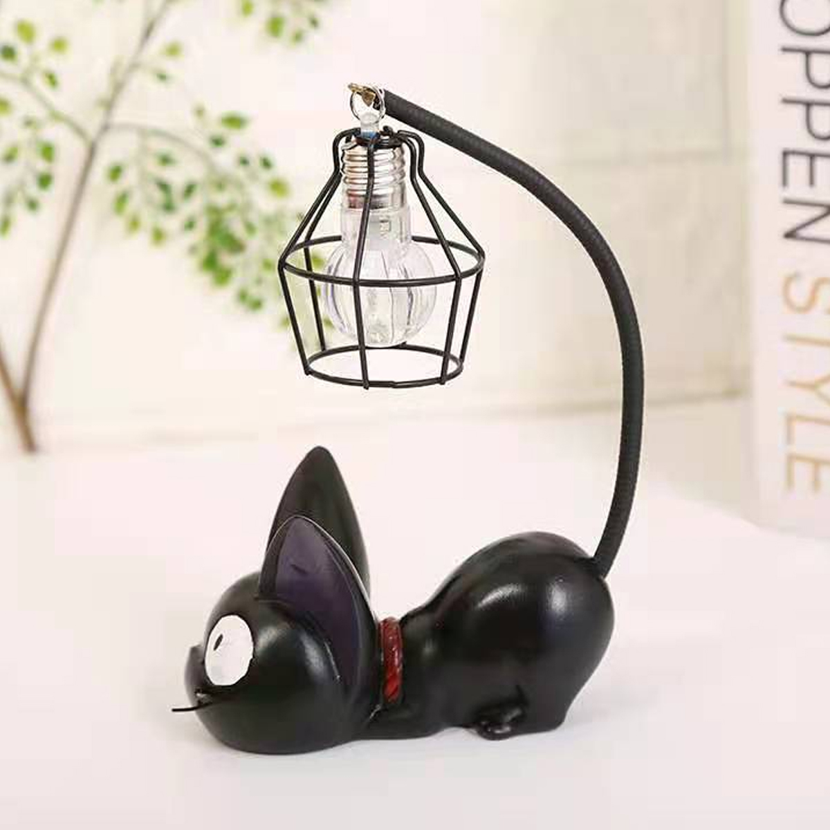 Eine schwarze Katzenförmige Lampe mit einer vergitterten Glühbirne. Die Katze liegt. Am Hals ist ein rotes Halsband.