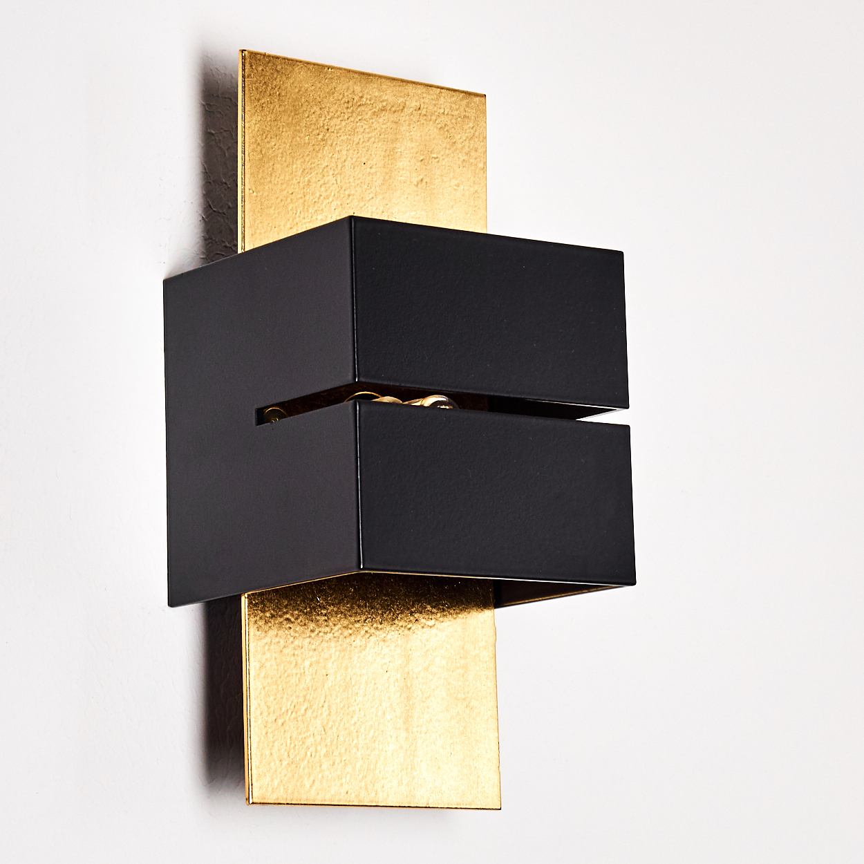 Eine moderne Wandleuchte mit einem schwarzen rechteckigen Rahmen vor einer goldfarbenen Platte an einer weißen Wand. Die Lampe hat einen Schlitz.