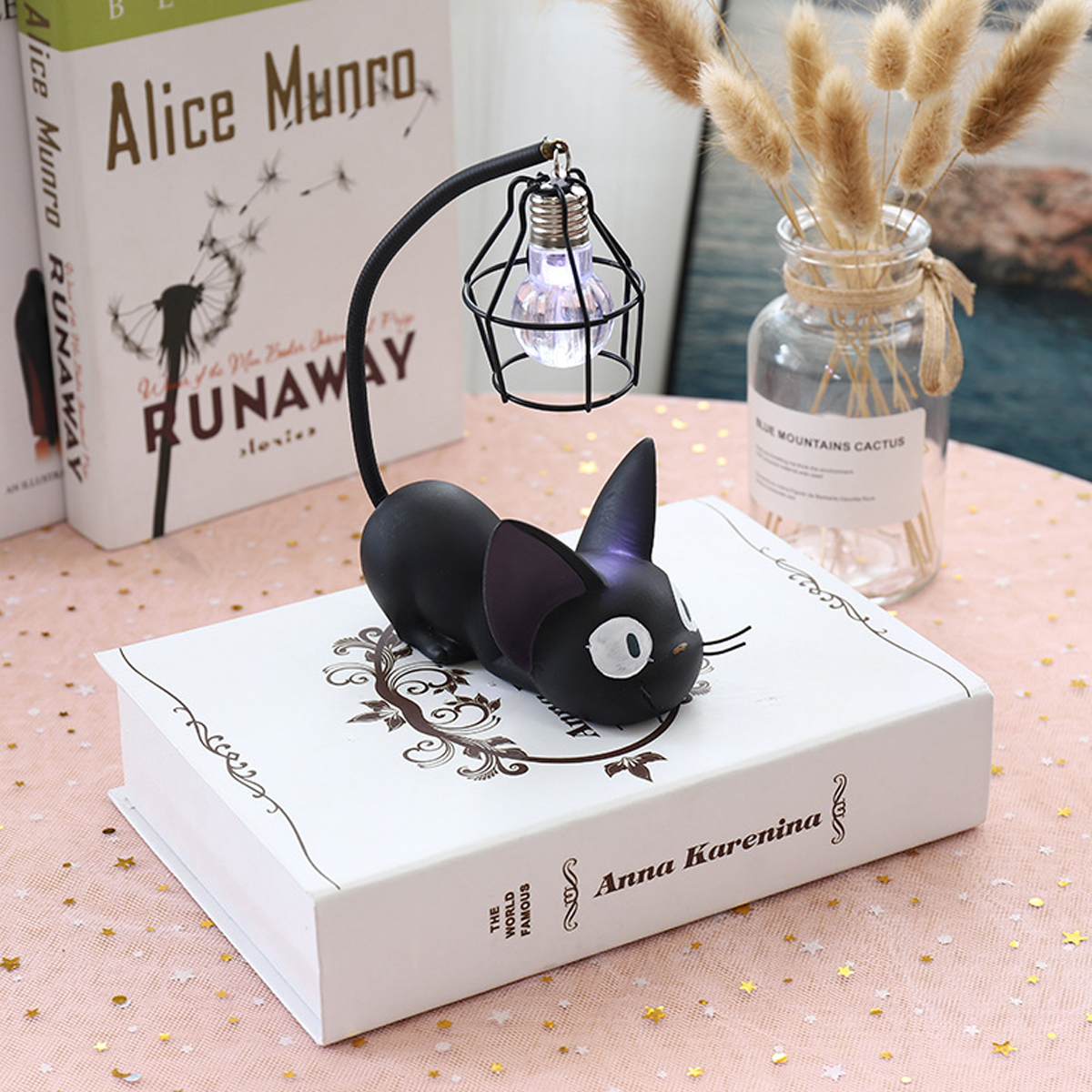 Eine schwarze Katzenlampe sitzt auf einem Buch. Über der Katze befindet sich eine Glühbirne. Bücher und eine Vase mit Pflanzen sind im Hintergrund.