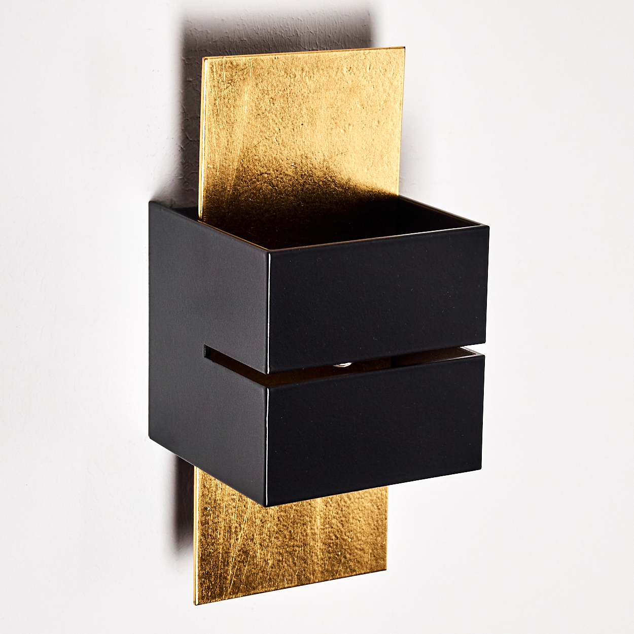 Eine moderne Wandleuchte. Sie hat einen schwarzen rechteckigen Kasten mit goldenem Blatt-Hintergrund. Die Leuchte ist an einer weißen Wand befestigt.