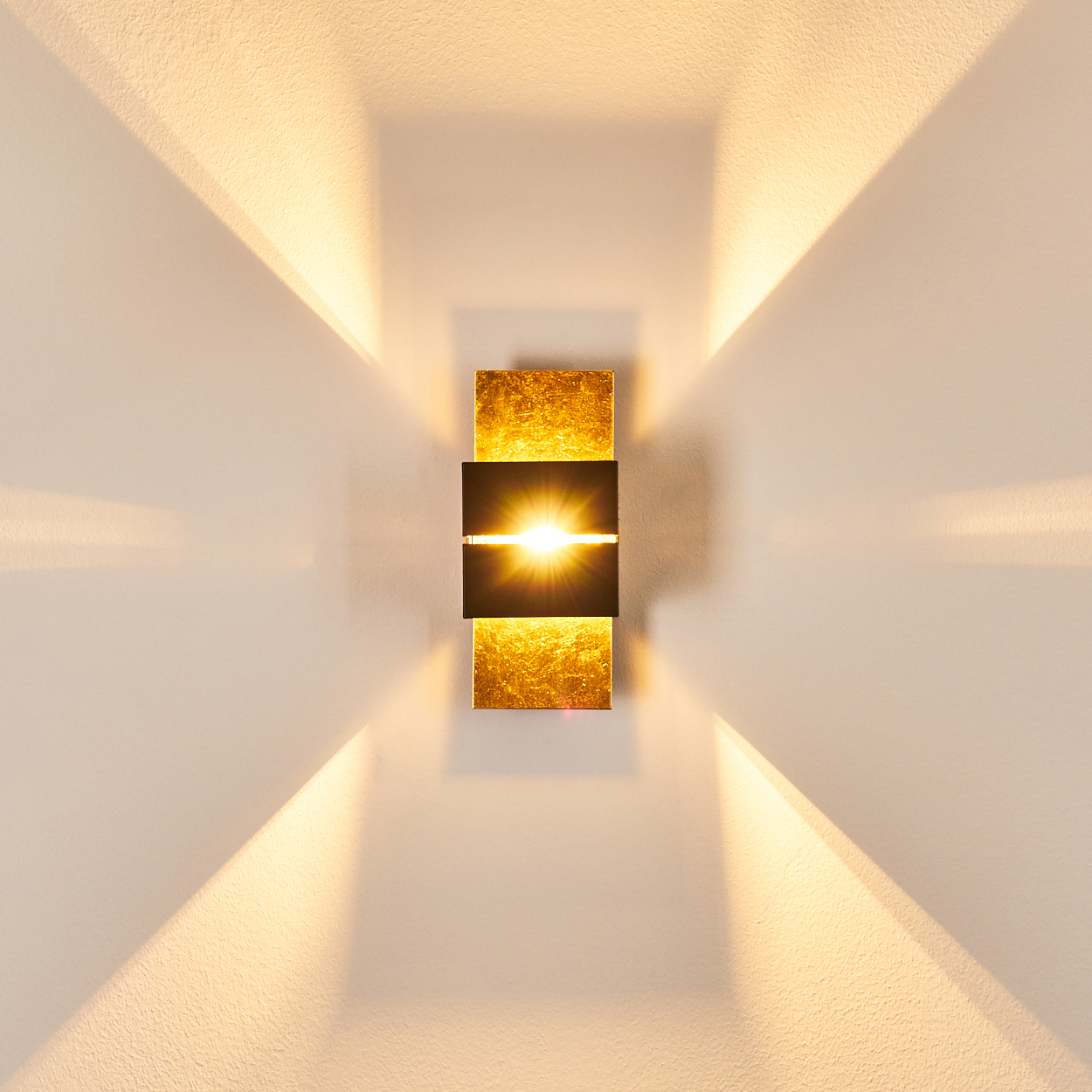 Eine Wandleuchte mit goldenen und schwarzen rechteckigen Elementen. Lichtstrahlen strahlen nach außen. Die Wand ist weiß.