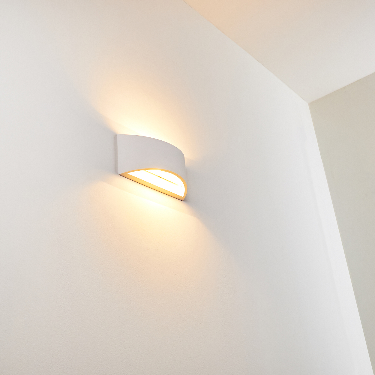 Eine weiße, an der Wand befestigte Leuchte mit gewölbtem Schirm. Sie strahlt warmes Licht aus und wirft einen Schatten an die Wand.