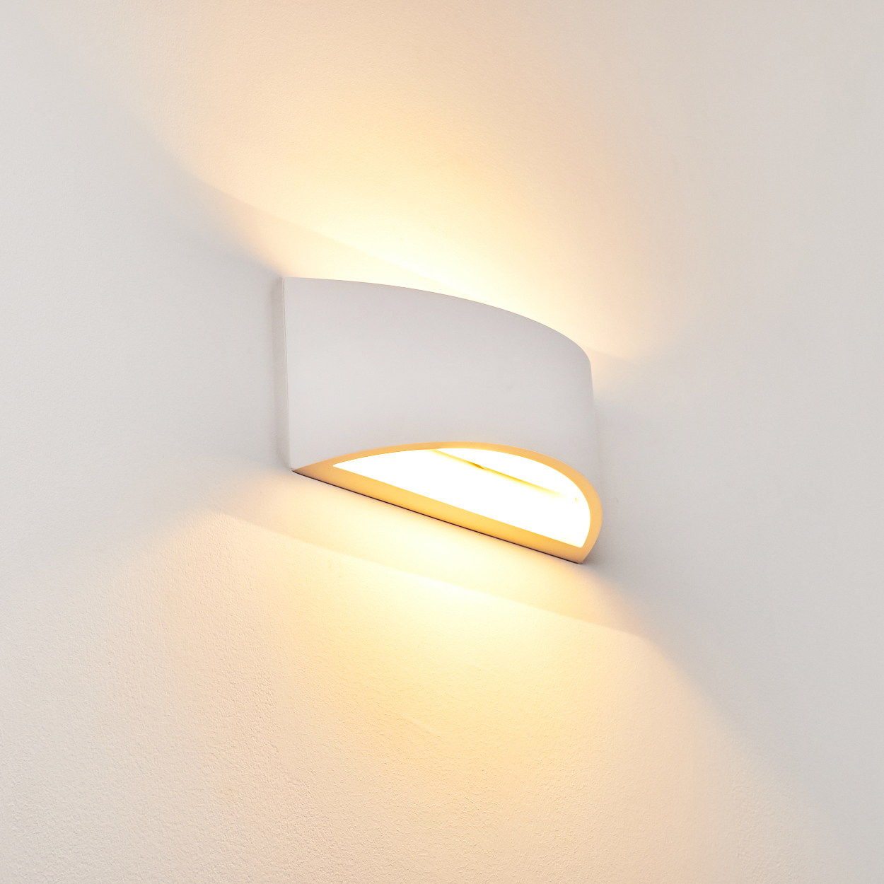 Eine weiße Wandleuchte mit geschwungenem Design an einer weißen Wand. Die Lampe strahlt warmes Licht aus und wirft Schatten.