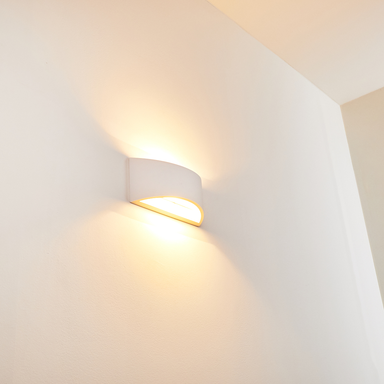 Eine weiße Wandleuchte gibt warmes Licht ab. Die Lampe ist gebogen, mit goldenem Innenraum, der das Licht nach oben und unten wirft.