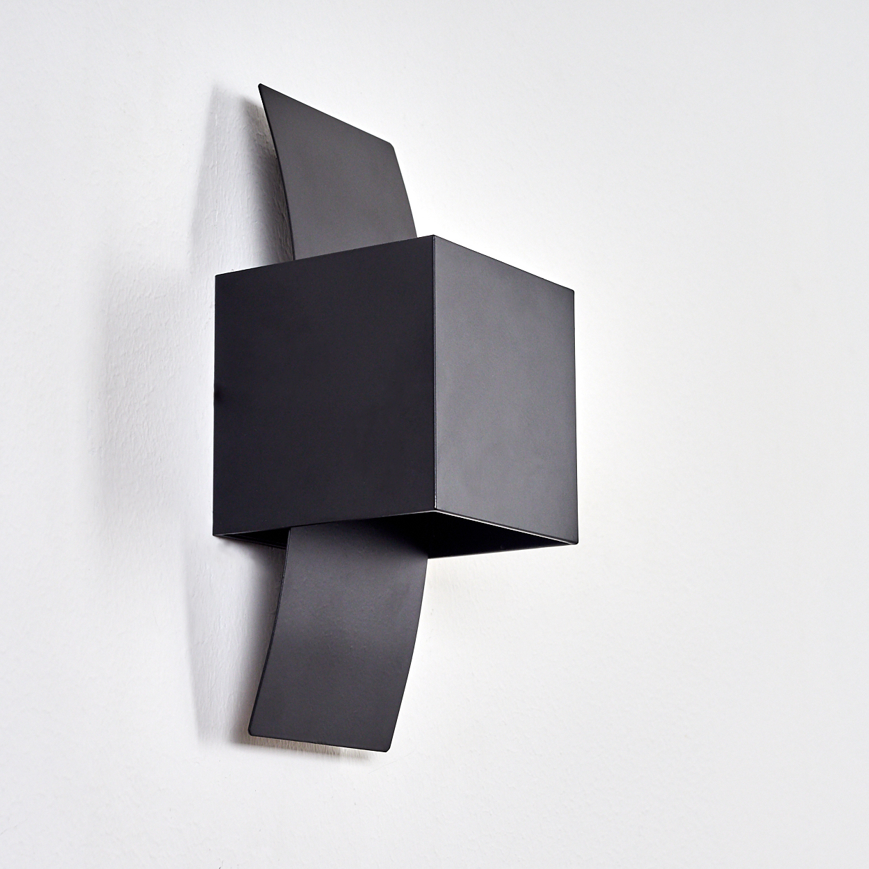 Schwarze, geometrische Wandleuchte. Es ist ein Würfel mit gebogenen rechteckigen Erweiterungen. An einer weißen strukturierten Wand montiert.