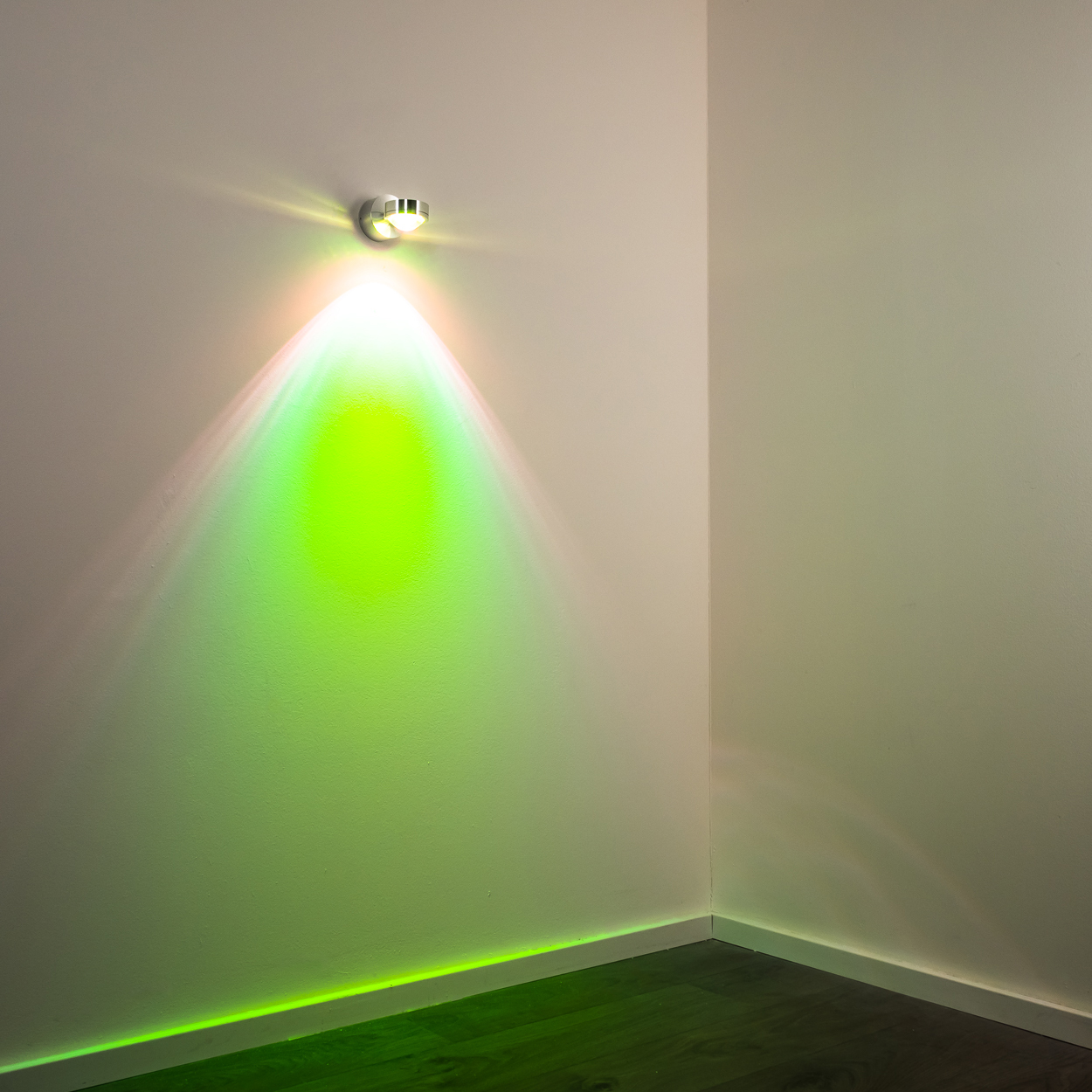 Ein Scheinwerfer an einer Wand beleuchtet eine Ecke. Grünes Licht breitet sich an der Wand aus, mit einer silbernen Halterung oben.