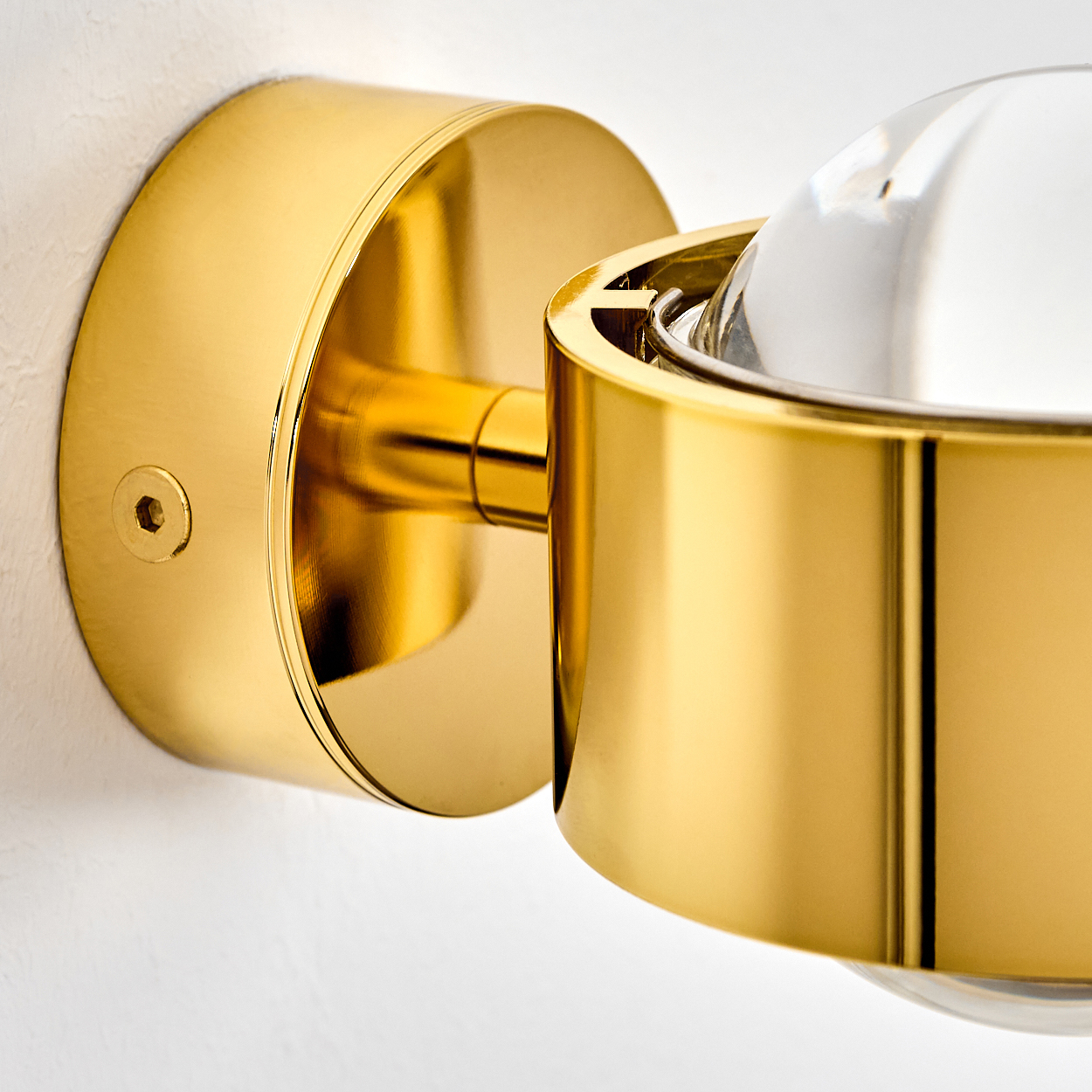 Nahaufnahme einer goldfarbenen Wandleuchte. Sie hat einen runden Sockel und eine klare Glaskugel. Die Lampe ist an einer weißen Wand befestigt.