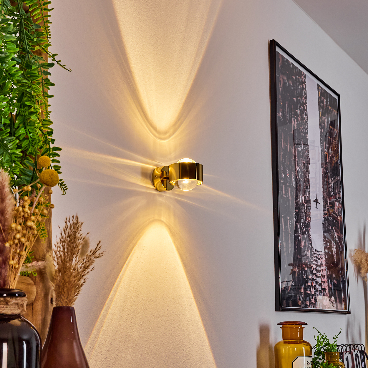 Eine goldene Wandlampe strahlt Licht auf eine weiße Wand. Rechts hängt ein Bild mit einer Stadtszene. Links befinden sich dekorative Pflanzen.