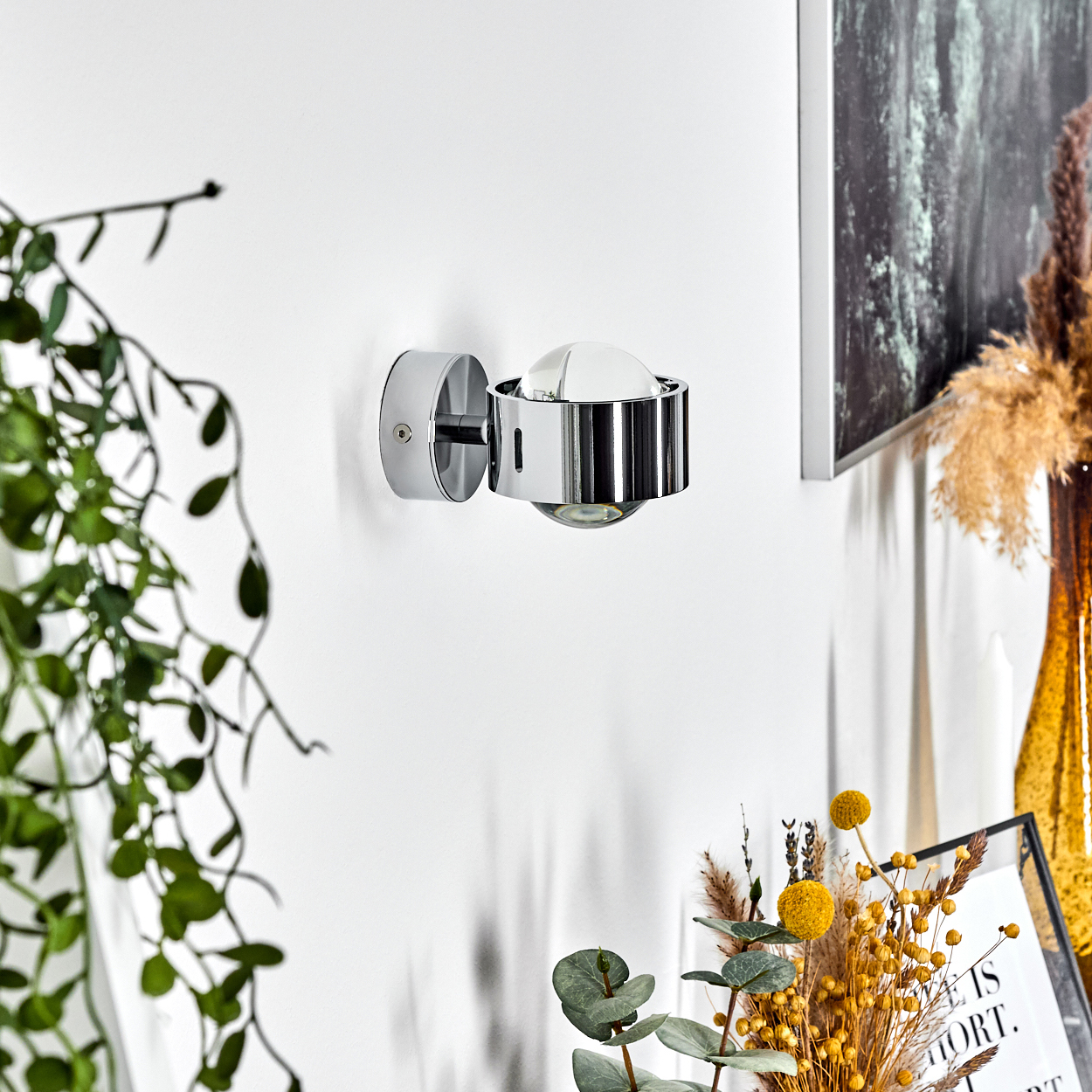 Eine verchromte Wandlampe ist an einer weißen Wand montiert. Grüne Pflanzen und Trockenblumen sind im Vordergrund. Ein Bilderrahmen ist ebenfalls zu sehen.