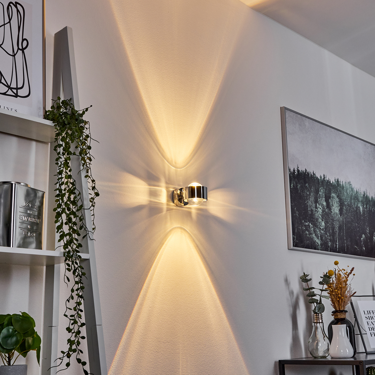 Wandleuchte, die nach oben und unten leuchtet. Weiße Wand. Dekoration umfasst Pflanze, gerahmte Kunst und dekorative Gegenstände.