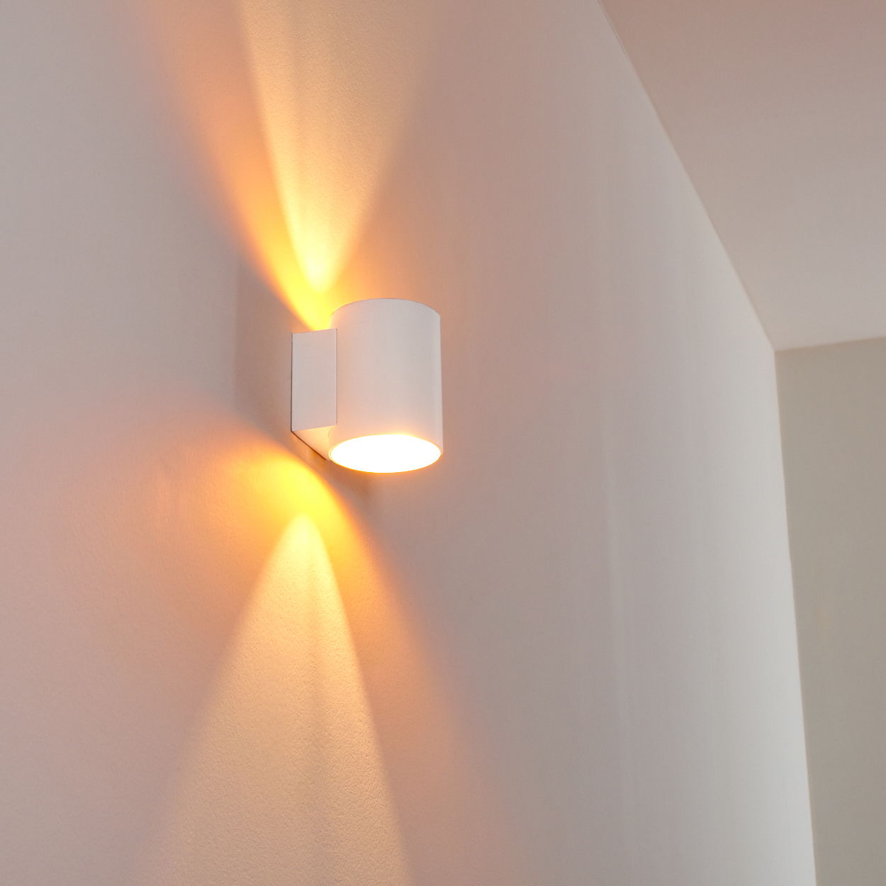 Eine weiße Wandleuchte strahlt nach oben und unten und wirft einen orangefarbenen Schein auf die weiße Wand. Die Lampe ist zylindrisch.