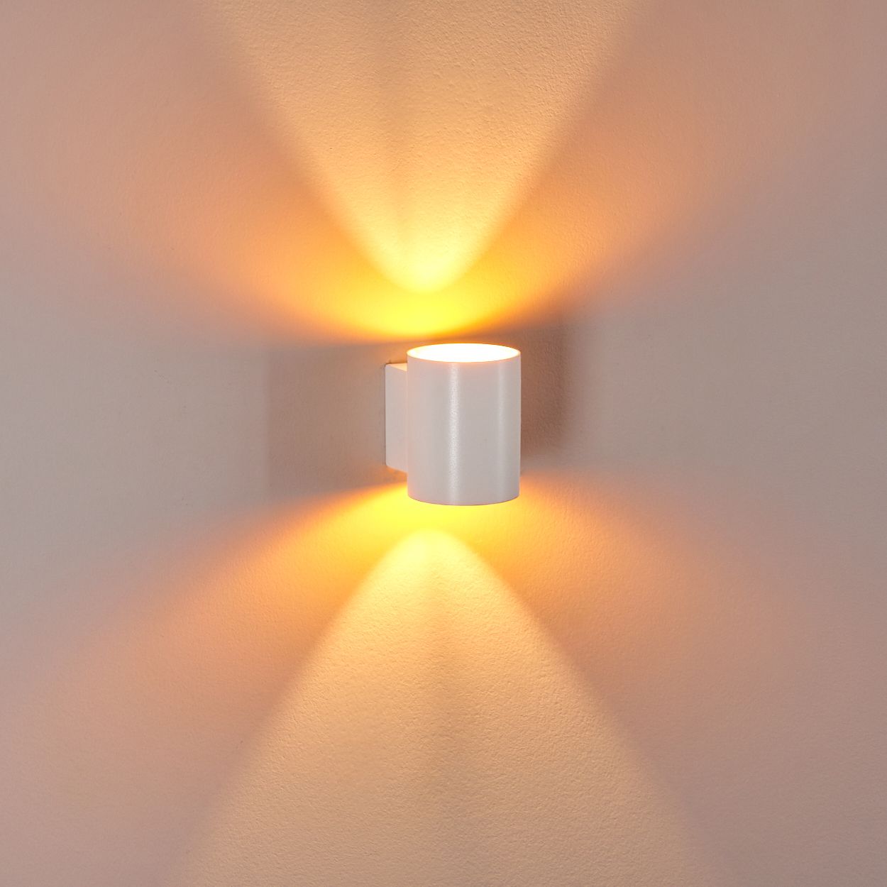 Eine weiße zylindrische Wandleuchte leuchtet und wirft orangefarbenes Licht auf die beige Wand. Das Licht erzeugt ein symmetrisches Muster.