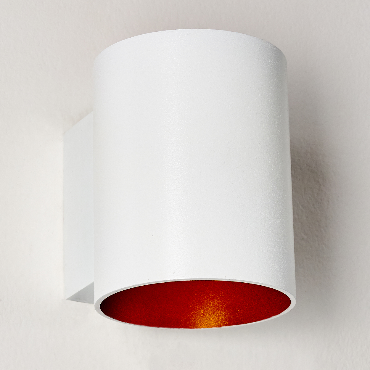 Eine weiße zylindrische Wandleuchte mit rotem Innenraum. Sie ist an einer weißen Wand montiert. Die Lampe hat ein schlichtes, modernes Design.