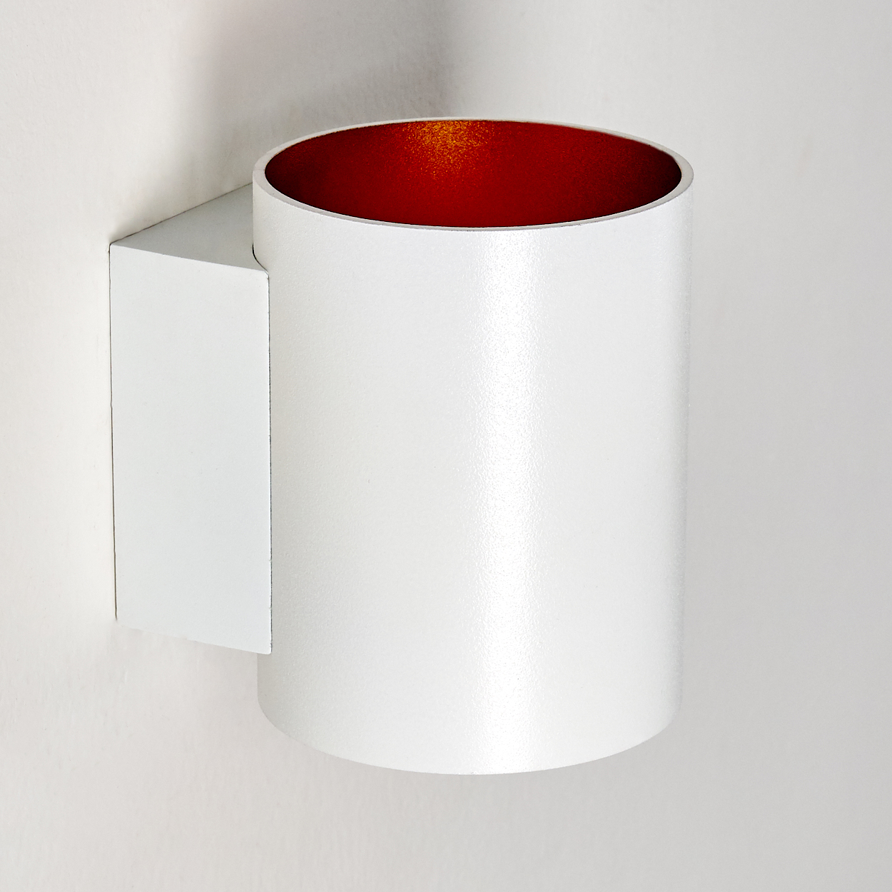 Eine weiße zylindrische Wandleuchte mit rotem Innenraum. Sie ist an einer weißen Wand montiert. Die Leuchte ist minimalistisch und modern.