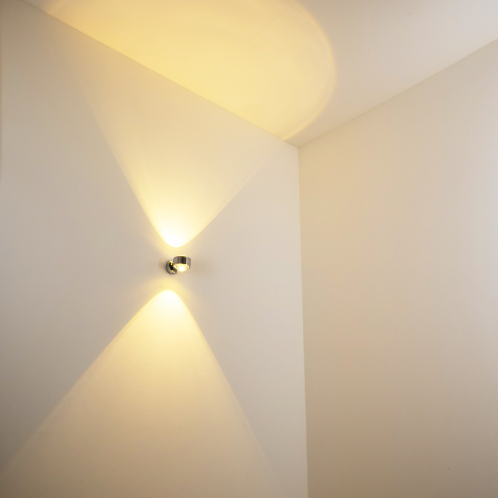 Eine moderne Wandleuchte mit zwei Lichtstrahlen, die die weiße Wand beleuchten. Die Strahlen erzeugen dreieckige Formen.