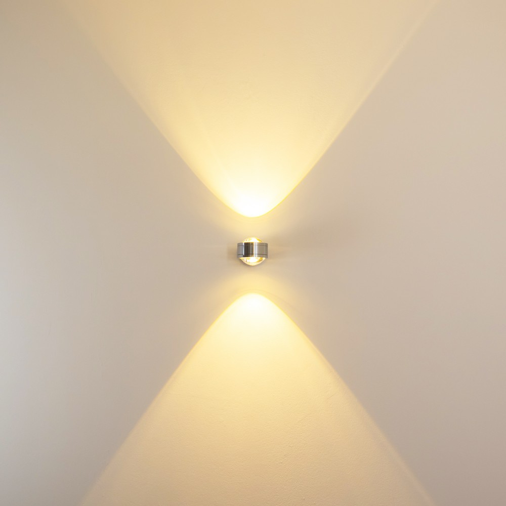 Eine moderne Wandleuchte ist an einer hellbeige Wand montiert. Die Lampe strahlt Licht nach oben und unten aus und erzeugt ein symmetrisches Lichtmuster.
