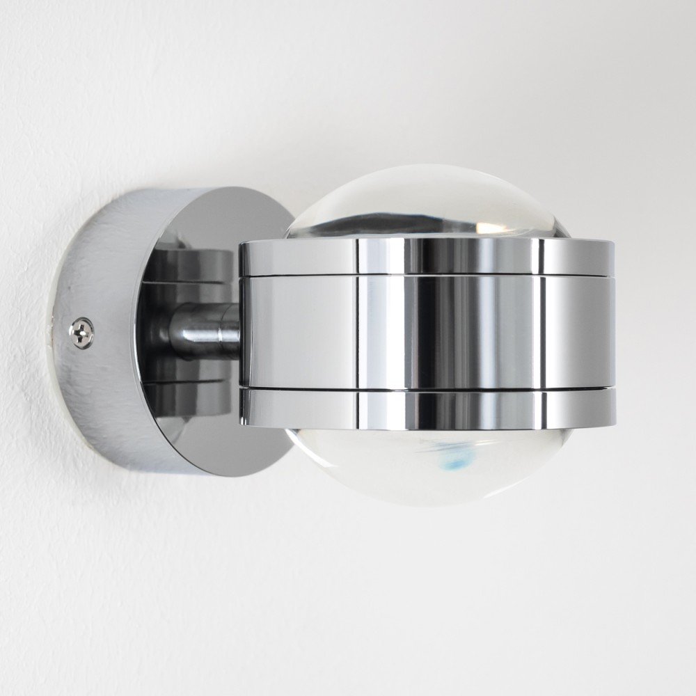 Eine verchromte Wandleuchte mit klarem, rundem Glasschirm. Sie befindet sich an einer weißen Wand. Die Lampe hat einen runden Sockel.