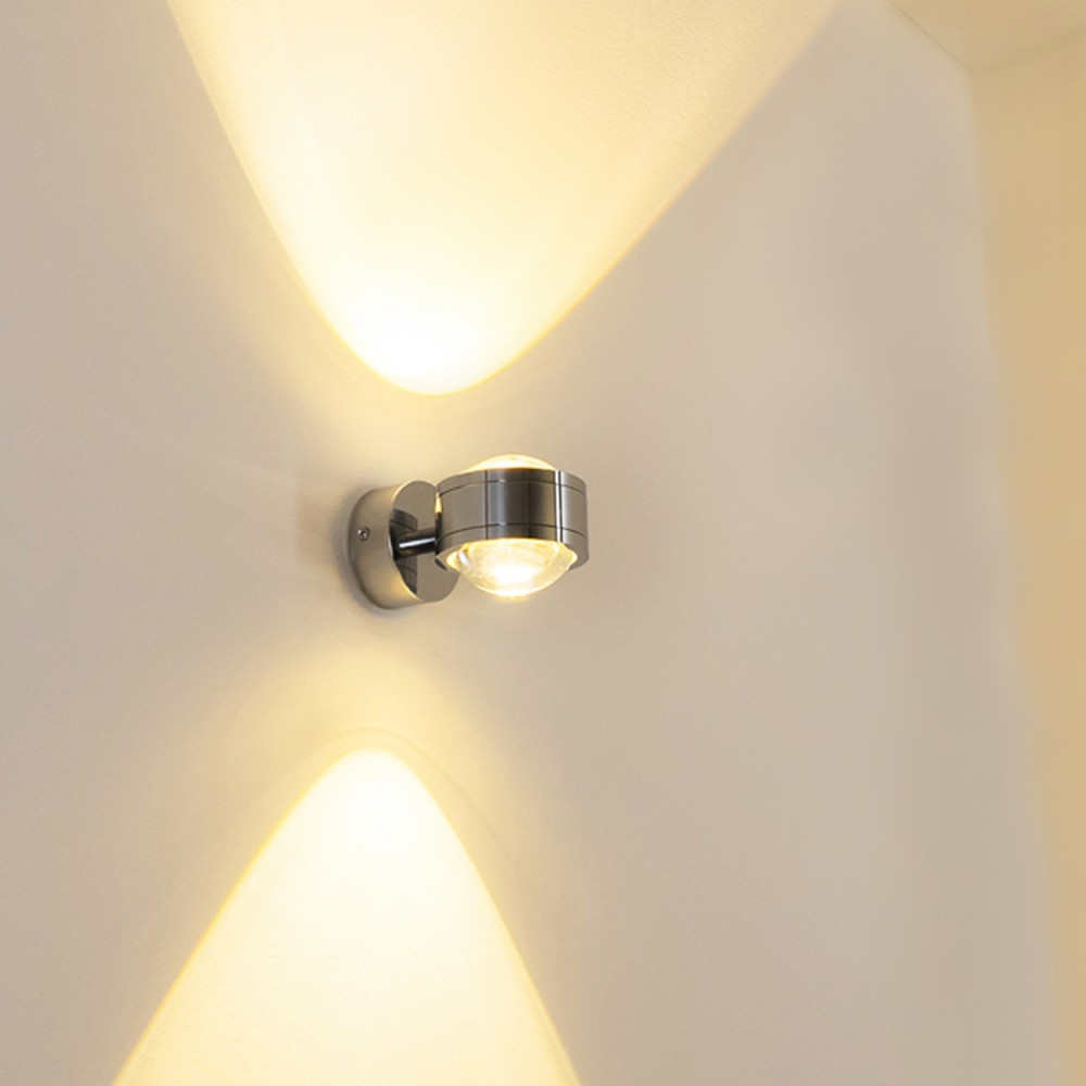 Eine moderne Wandleuchte mit Metalloberfläche ist an einer weißen Wand befestigt. Die Lampe strahlt helles Licht nach oben und unten aus.
