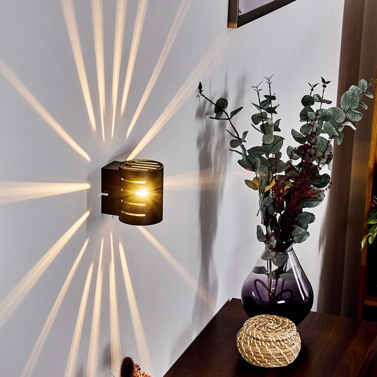Eine an der Wand montierte schwarze Lampe wirft Lichtmuster auf die Wand. Eine Vase mit Grün steht auf einem Holztisch. Ein geflochtener Korb ist vorhanden.