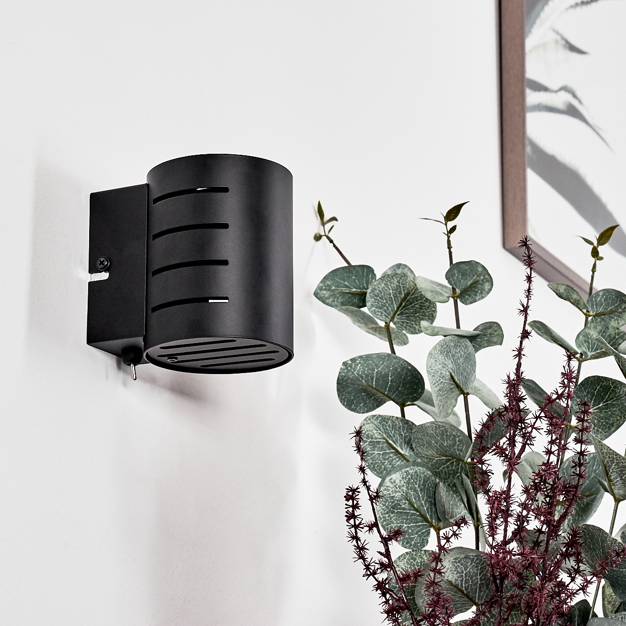 Schwarze Wandlampe mit Schlitzen, Eukalyptuspflanze, gerahmtes Bild an weißer Wand.