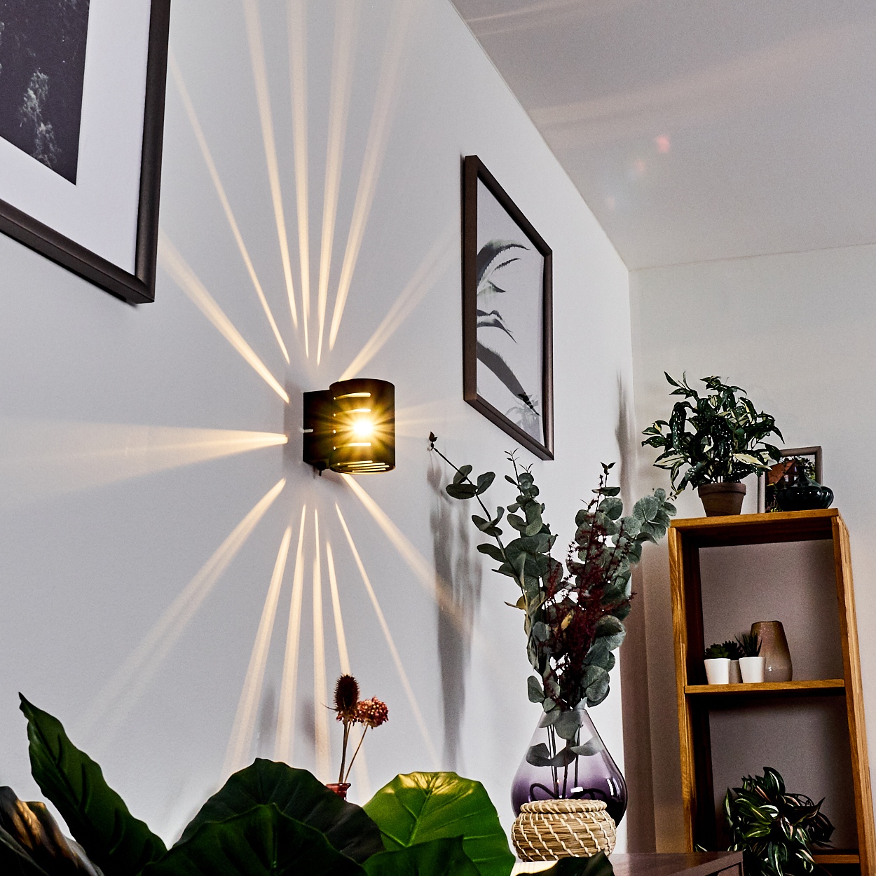 Eine moderne Wandlampe wirft Lichtstrahlen auf eine weiße Wand. Kunstwerke und Pflanzen schmücken den Raum und schaffen eine gemütliche Atmosphäre.