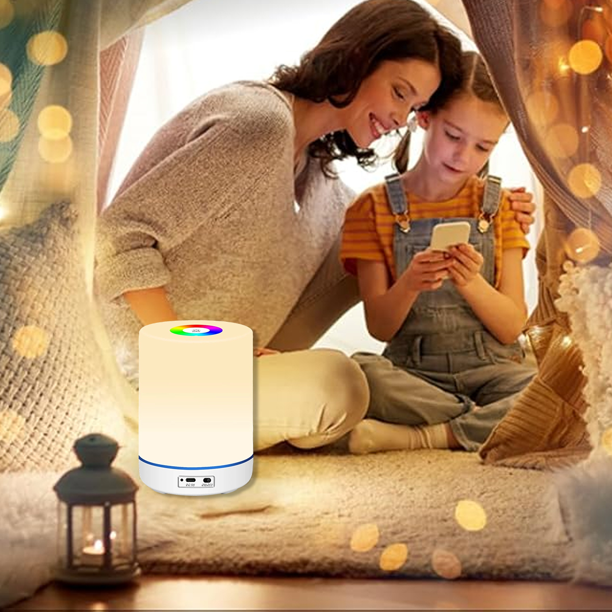 Una donna e una bambina sono sedute in una tenda, guardando un telefono. Una lampada e una lanterna sono davanti a loro. Scena accogliente con luci.
