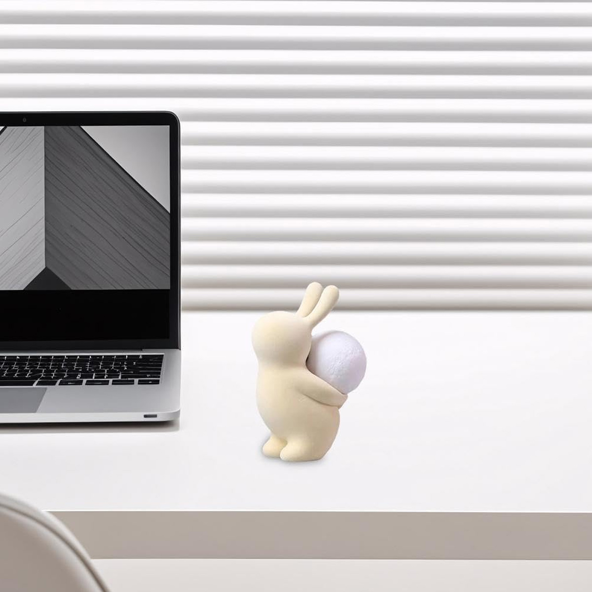 Eine beigefarbene Hasenfigur, die einen weißen Ball hält, sitzt auf einem weißen Schreibtisch neben einem Laptop. Der Hintergrund ist ein weißer Rollladen.