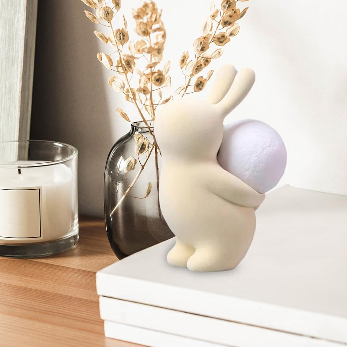 Eine cremefarbene Hasenfigur hält eine weiße Kugel. Im Hintergrund eine Kerze und eine Vase mit getrockneten Blumen. Der Hase sitzt auf Büchern.