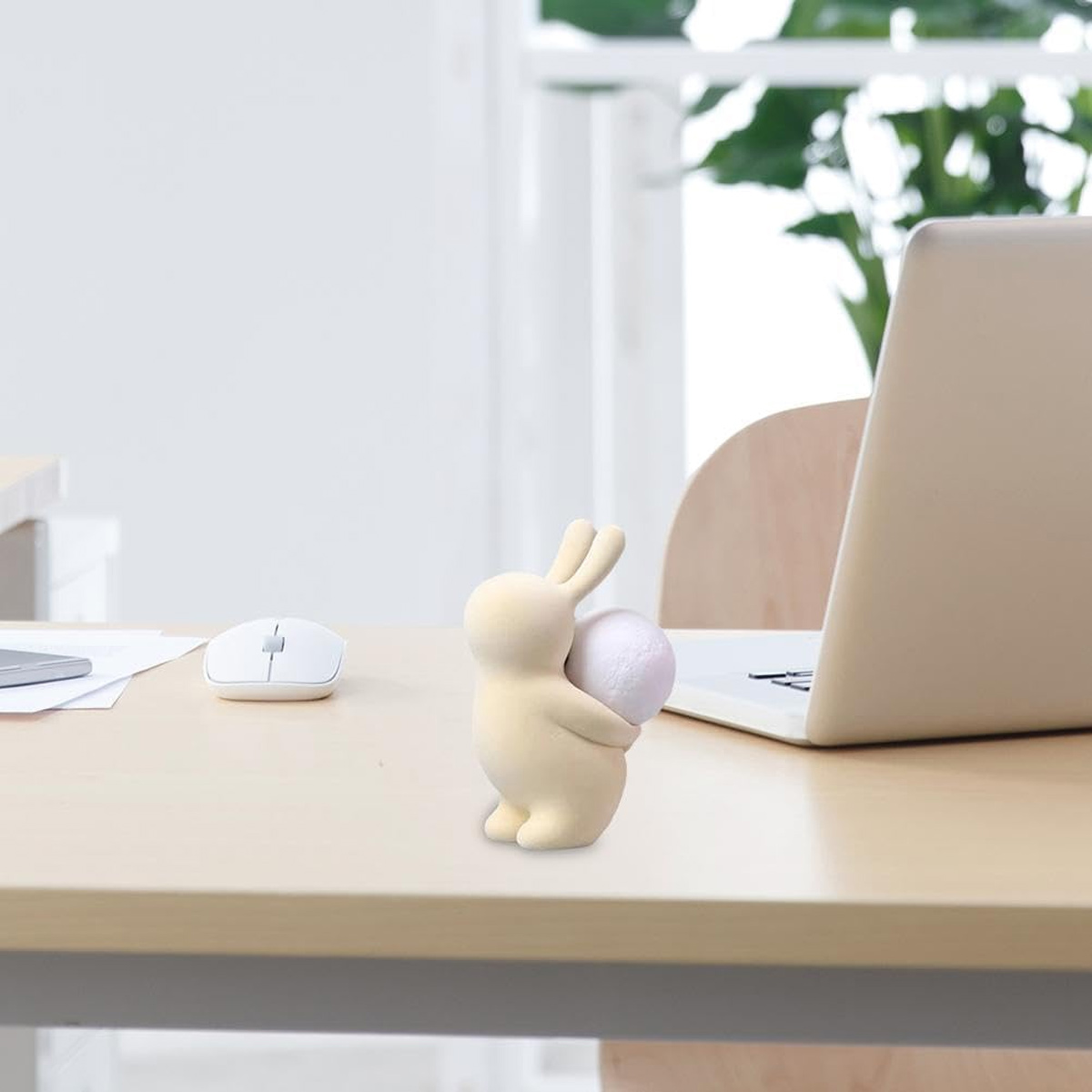 Eine beige Hasenfigur hält einen weißen Ball auf einem Schreibtisch mit Laptop und Maus. Weiches Sonnenlicht scheint durch ein Fenster.