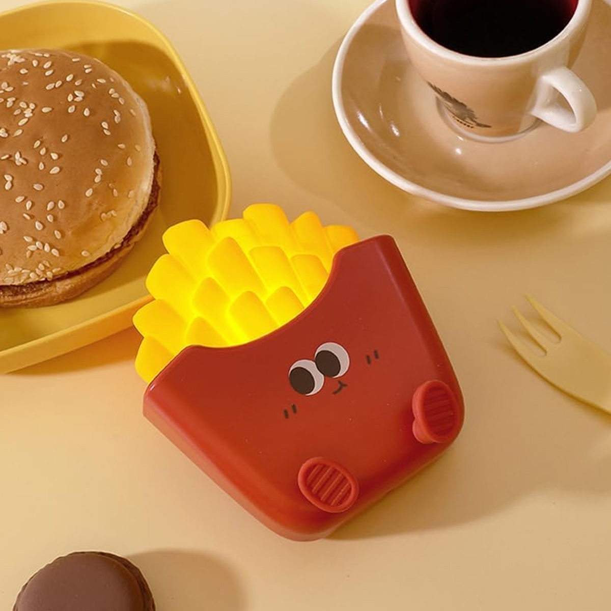 Eine rote Pommes-Lampe mit Smiley-Gesicht, Burger, Kaffee und gelber Gabel auf gelber Oberfläche.