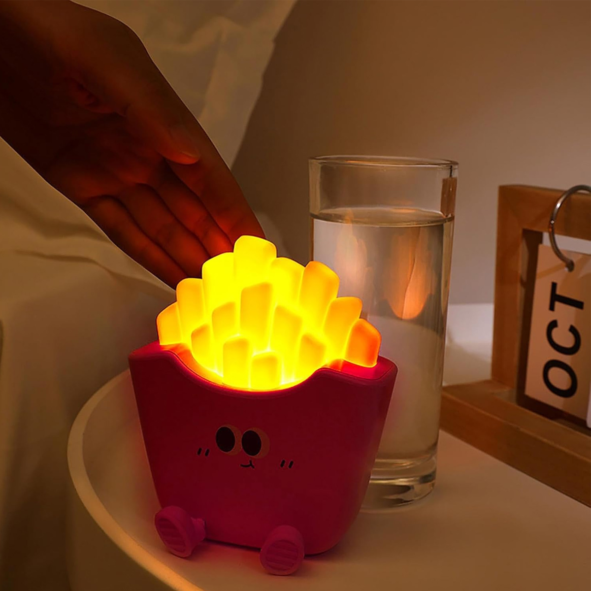 Eine Hand schwebt über einer Pommes-Lampe auf einem Nachttisch. Ein Glas Wasser und ein Oktoberkalender sind ebenfalls vorhanden.