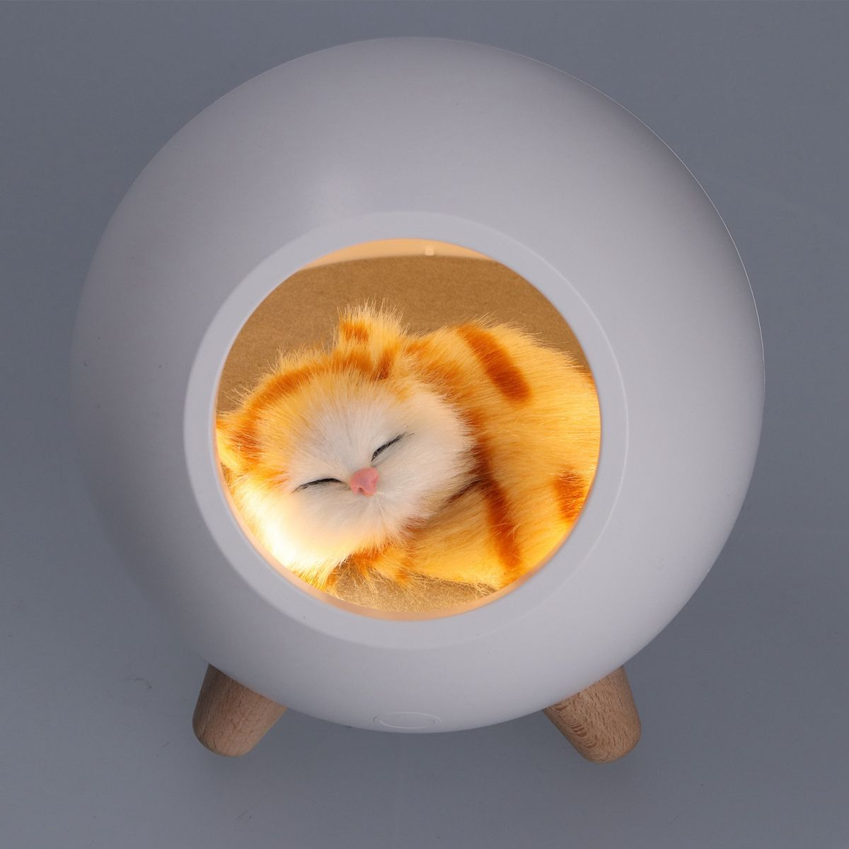 Ein schlafendes orange-weißes Katzenspielzeug in einem weißen kugelförmigen Bett mit Holzbeinen, von innen beleuchtet.
