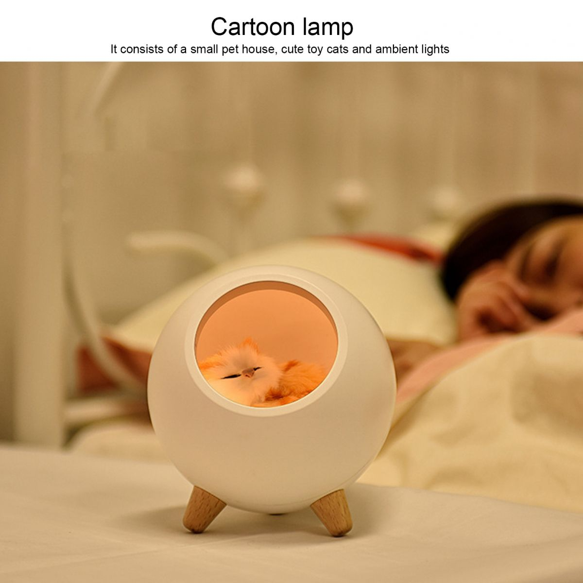 Eine weiße, kugelförmige Lampe mit einer Spielzeugkatze darin, die auf einem Bett liegt. Im Hintergrund schläft eine Person.