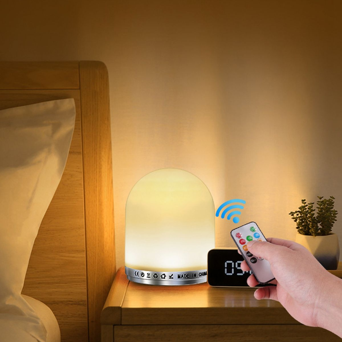 Eine Nachttischlampe mit Fernbedienung auf einem Nachttisch. Eine Hand zeigt mit der Fernbedienung auf die Lampe. Daneben eine Digitaluhr und eine Pflanze.