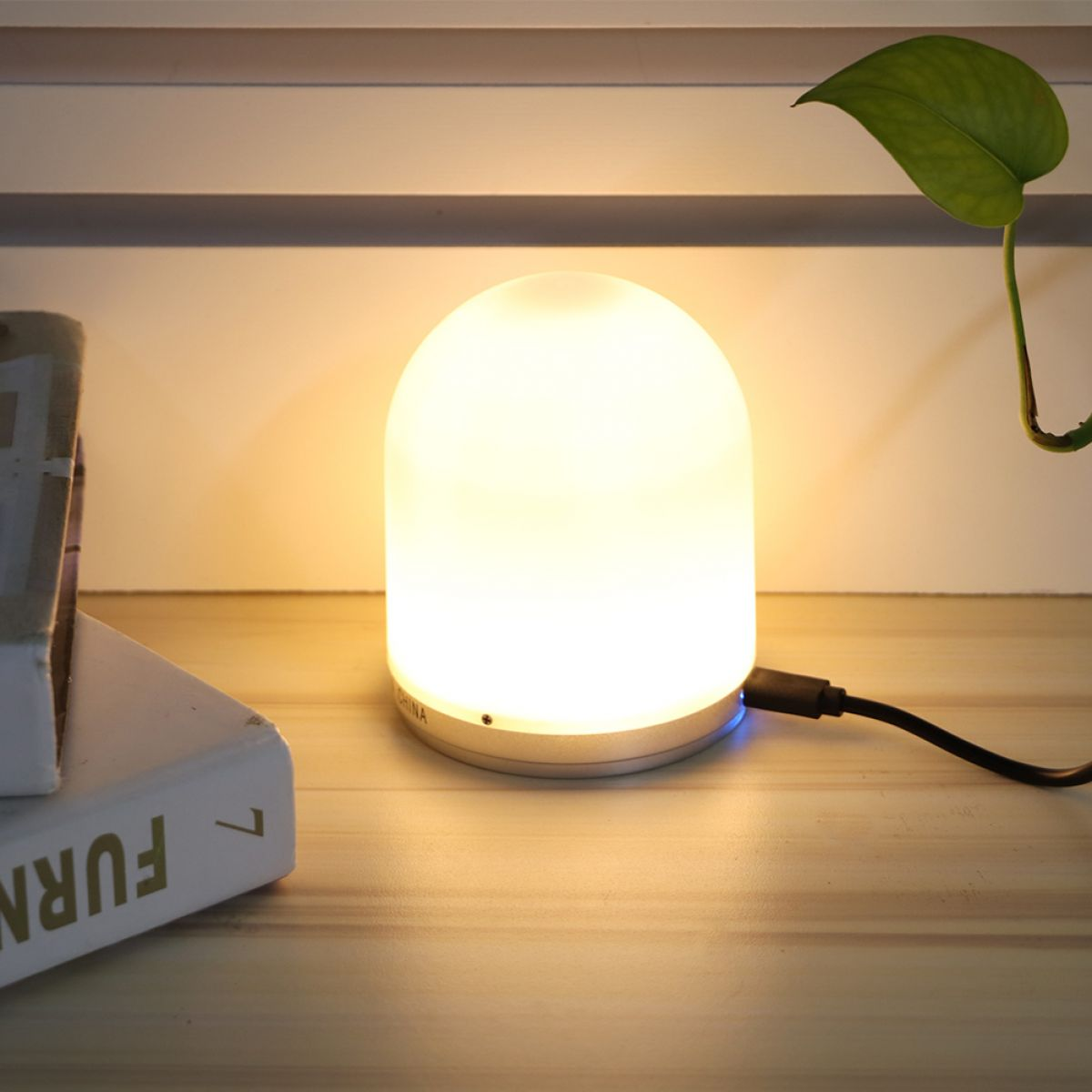Eine leuchtende weiße Lampe steht auf einer Holzoberfläche. Ein Buch und ein grünes Blatt sind ebenfalls zu sehen. Ein USB-Kabel ist an der Lampe angeschlossen.