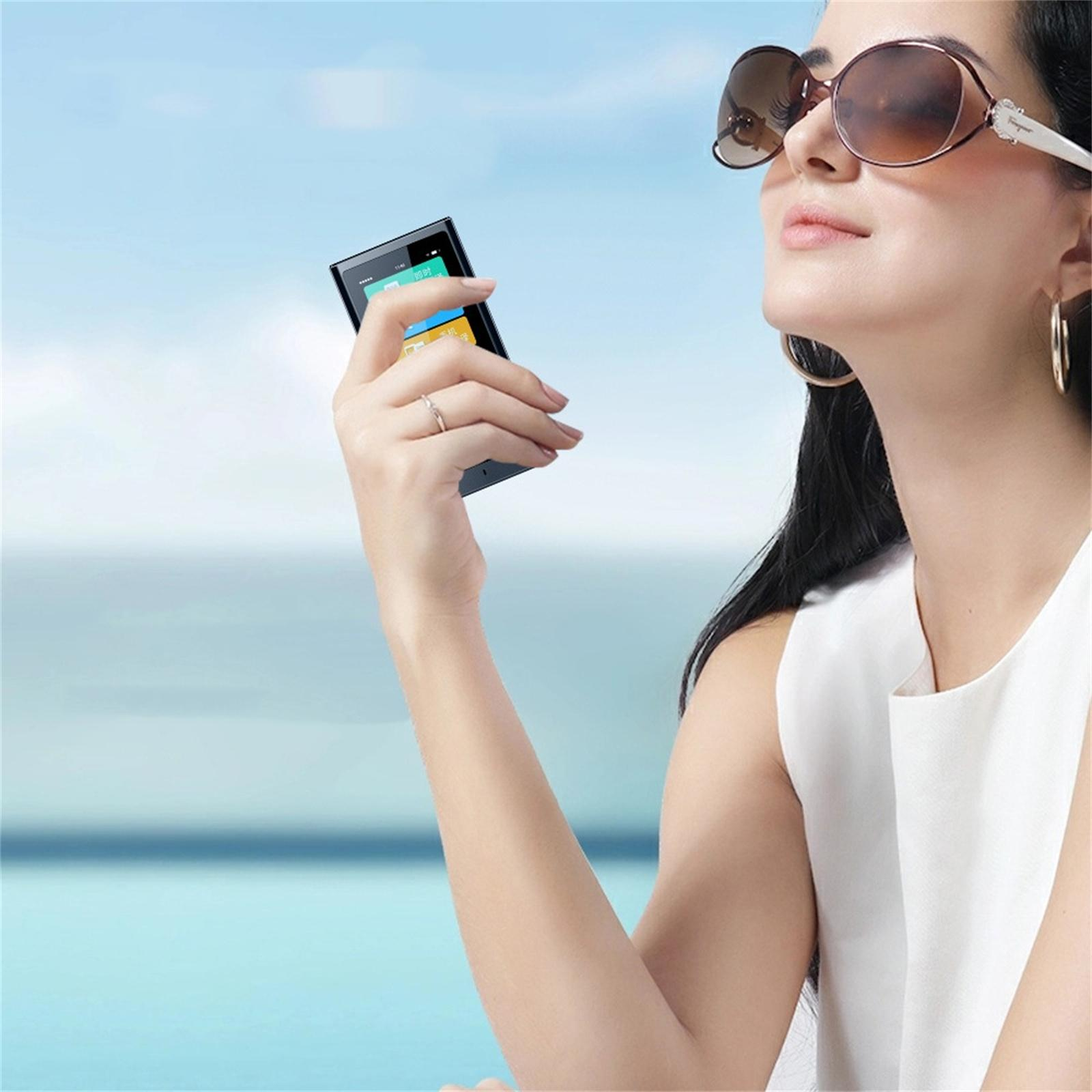 Frau mit Sonnenbrille hält ein Telefon. Blauer Himmel und Meer im Hintergrund. Sie trägt einen Ring und ein weißes Oberteil.