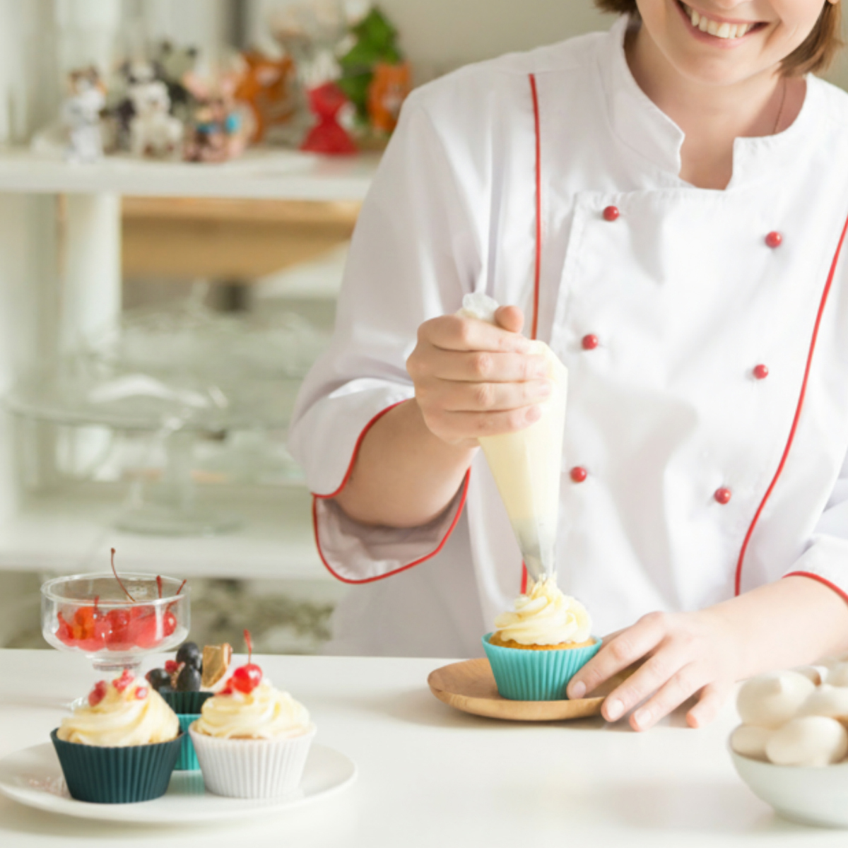 Eine Bäckerin in weißer Uniform mit rotem Besatz verziert einen Cupcake mit Sahne. Cupcakes und Kirschen stehen auf dem Tisch.