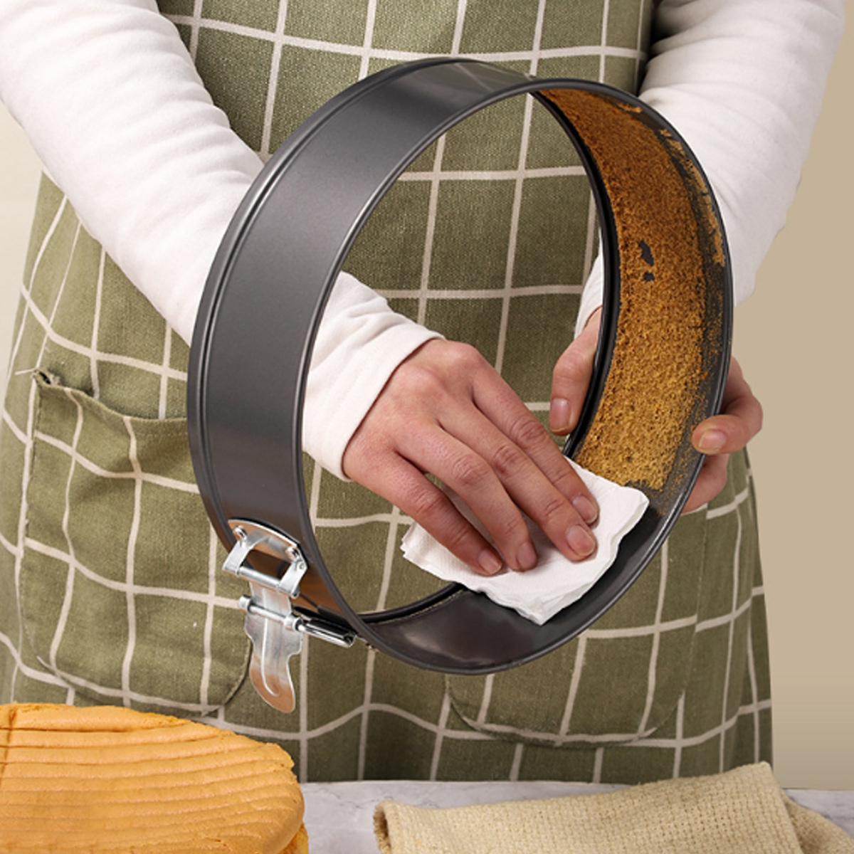 Eine Person mit Schürze reinigt eine Kuchenform mit einem weißen Tuch. Ein Kuchen und ein Tuch sind im Vordergrund.