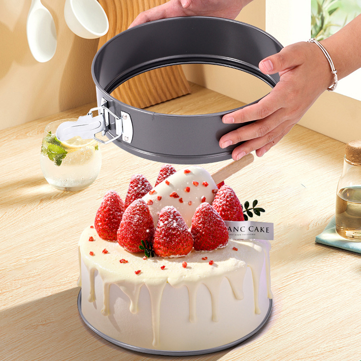 Ein Kuchen mit Erdbeeren und einem Eis am Stiel im Vordergrund. Eine Person hält eine Kuchenform darüber. Ein Getränk ist links.
