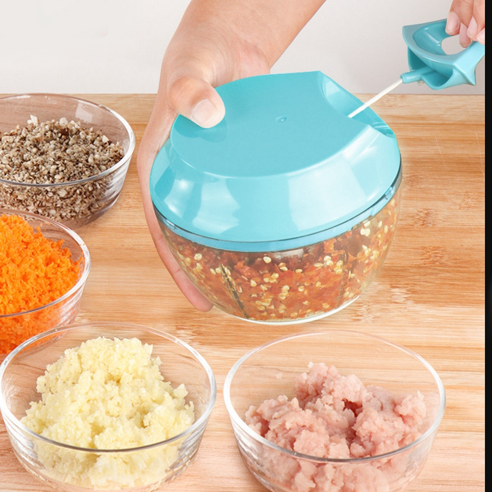 Eine Hand hält einen blauen Zerkleinerer über einer Holzoberfläche. Es gibt Schalen mit gehackten Lebensmitteln: Karotten, Knoblauch, Fleisch und andere Zutaten.