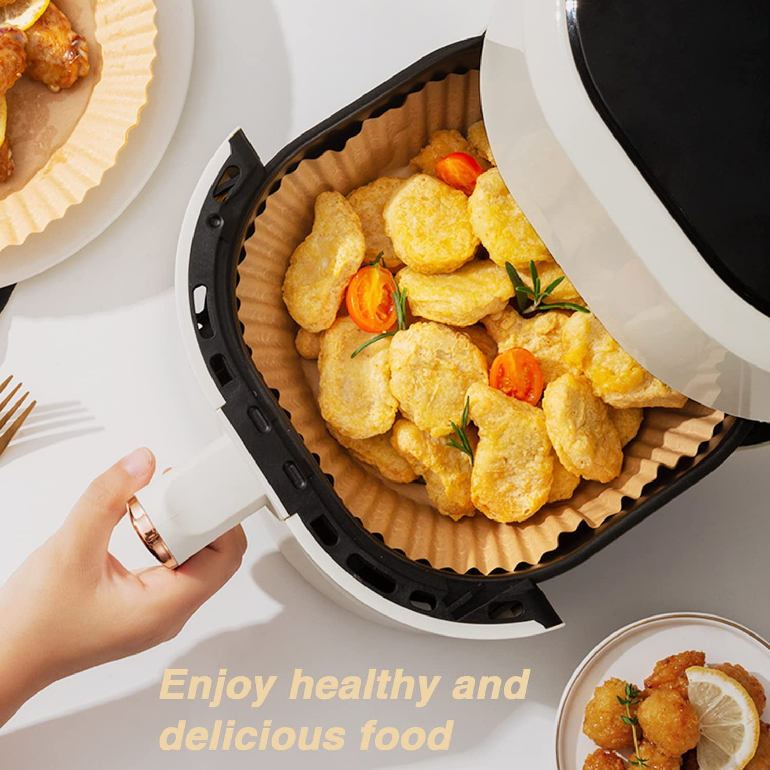 Ein Airfryer mit Nuggets, Tomaten und Kräutern. Eine Hand hält den Griff. Teller mit gebratenem Essen. Text: Genieße gesundes und leckeres Essen.