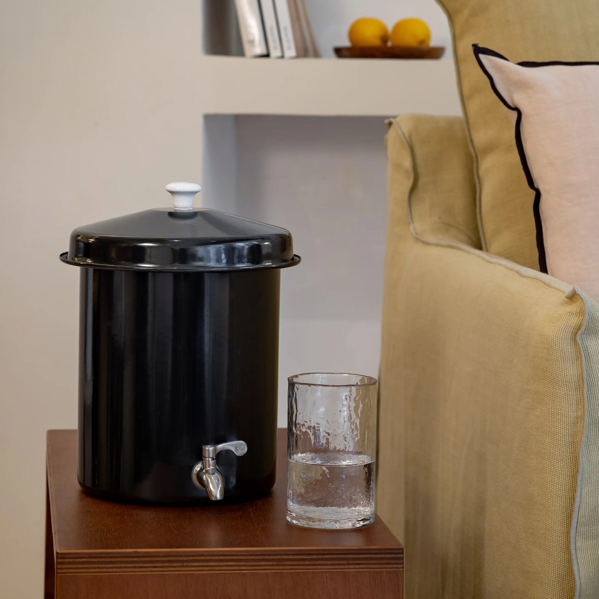 Un distributore d'acqua nero, bicchiere d'acqua su un tavolo di legno. Divano e limoni sullo sfondo.
