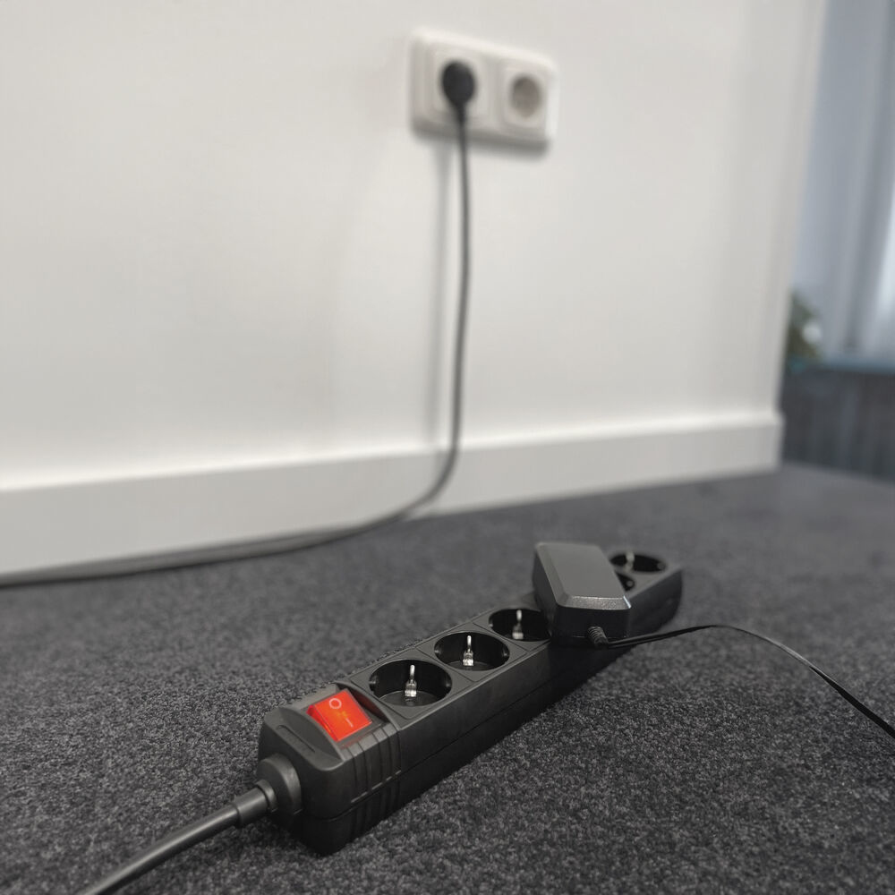 Une multiprise noire avec un interrupteur rouge est sur un tapis gris. Un mur blanc avec une prise est en arrière-plan.