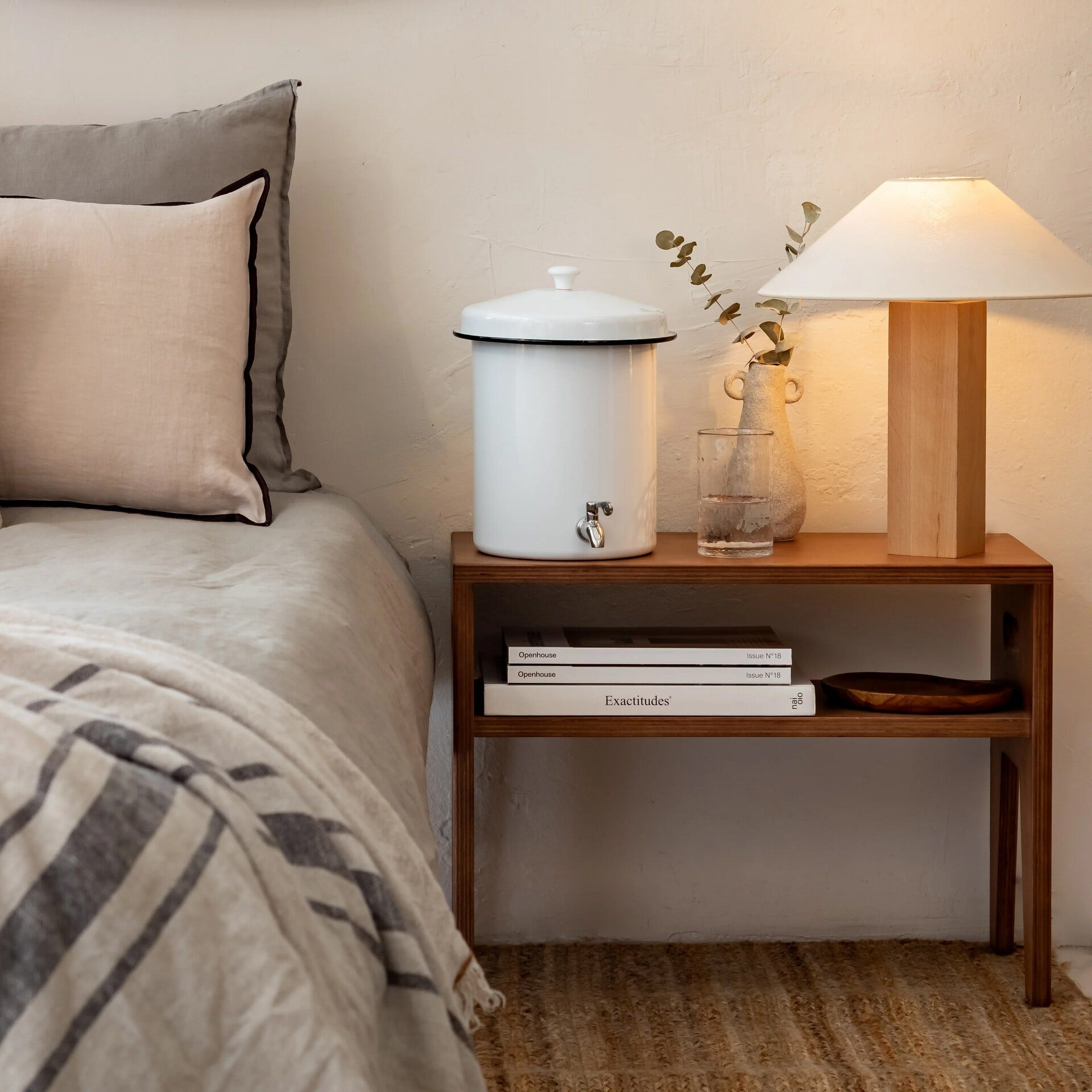 Comodino con lampada, distributore d'acqua, libri e vaso. Letto e tappeto sullo sfondo.